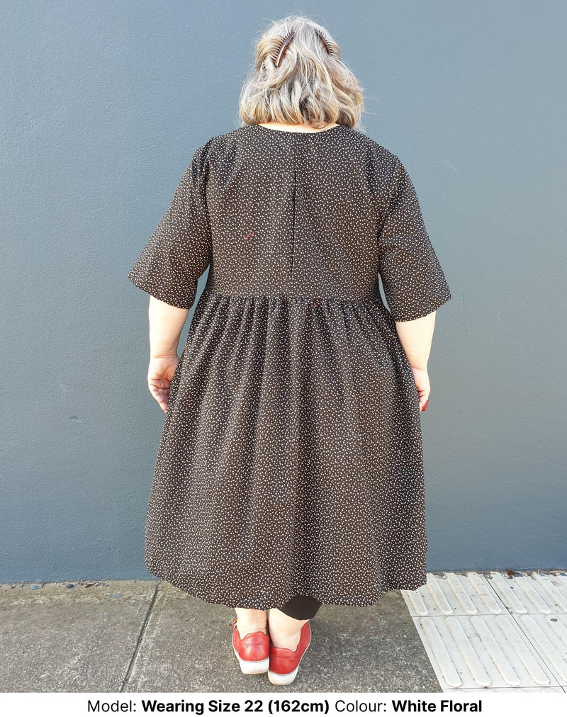 Back view of Model wearing size 22 plus size midi dress in black cotton with small floral motif showing pleat at centre back