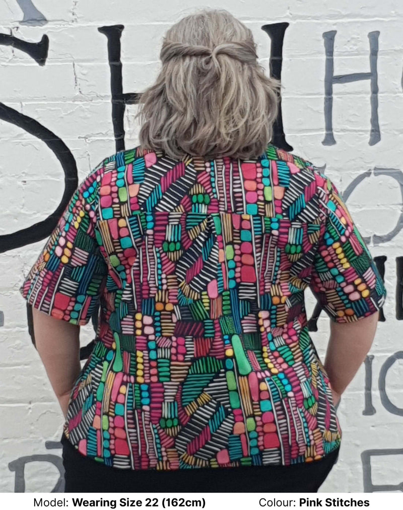 back view of Bright Watercolour print blouse in plus sizes. The print is predominately green, pink, white, blue and yellow and looks very cheerful against a black background. The blouse is by Chasing Springtime and the model is wearing size 22
